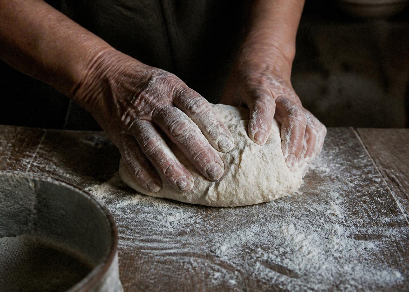 Weathered hands working bread dough on a floured wooden table.